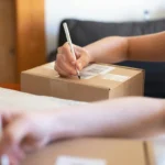 Tangan seseorang menulis label pada kardus paket untuk keperluan pengiriman dari rumah